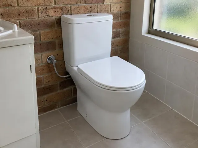 White close-coupled toilet beside a brick wall and tiled floor near a window in Lakelands
